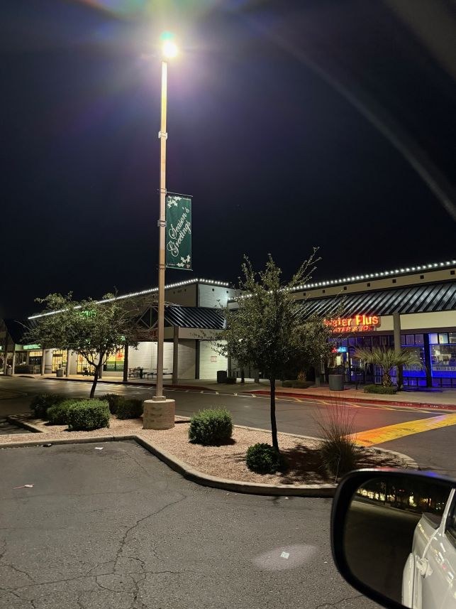 A green Season’s Greetings holiday banner on a parking-lot light pole with roofline lights behind.