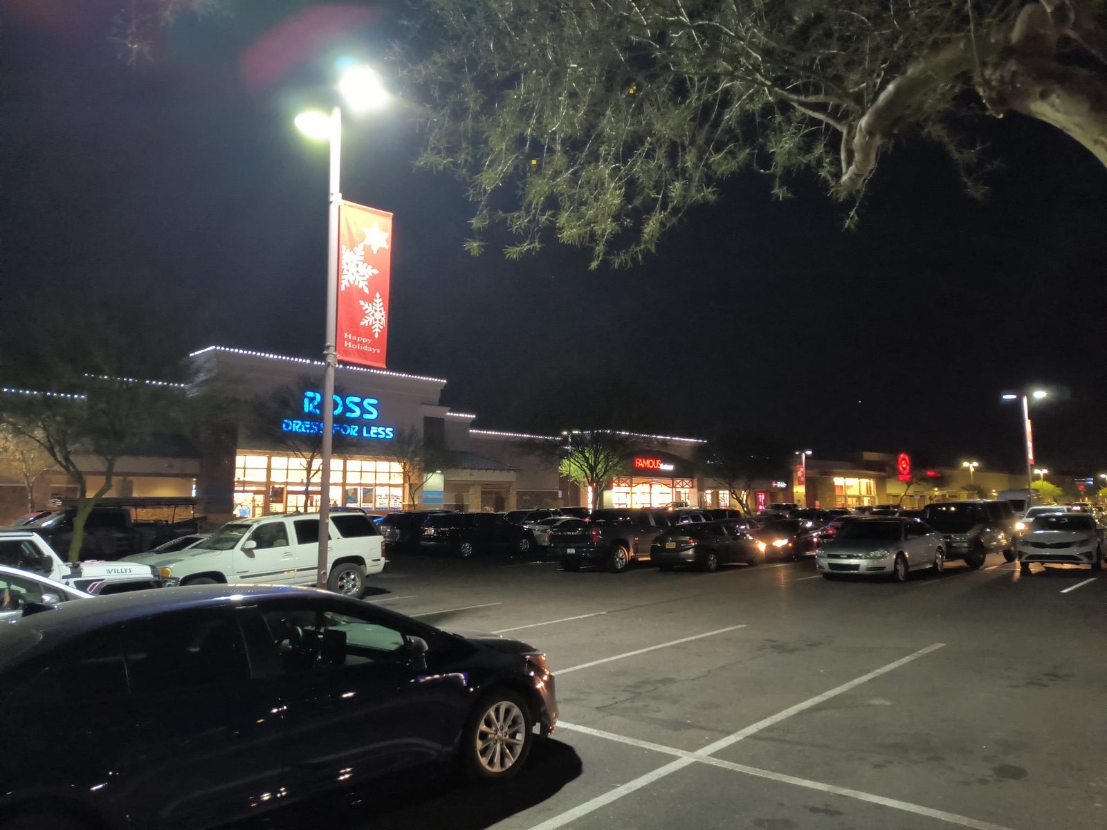 A red Happy Holidays snowflake banner mounted on a parking-lot light pole, with C9 roofline lights across the shopping center behind it.